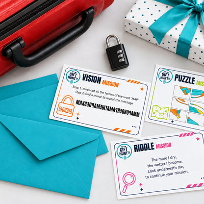 A flat-lay of gift puzzle cards, teal envelopes, a small black padlock, a red suitcase, and a wrapped gift box on a white surface. The cards show mission prompts such as Vision Mission, Puzzle Mission, and Riddle Mission, with colorful graphics and text.