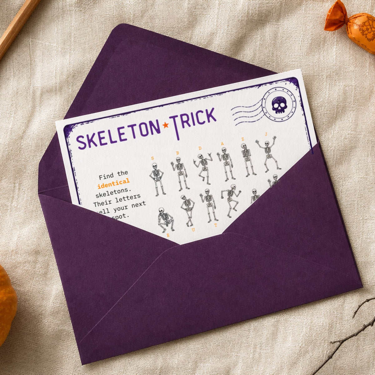 A Halloween-themed postcard labeled 'SKELETON TRICK' tucked inside a dark purple envelope, styled on a beige linen fabric background with small orange decorative accents.