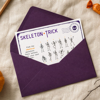 A Halloween-themed postcard labeled 'SKELETON TRICK' tucked inside a dark purple envelope, styled on a beige linen fabric background with small orange decorative accents.