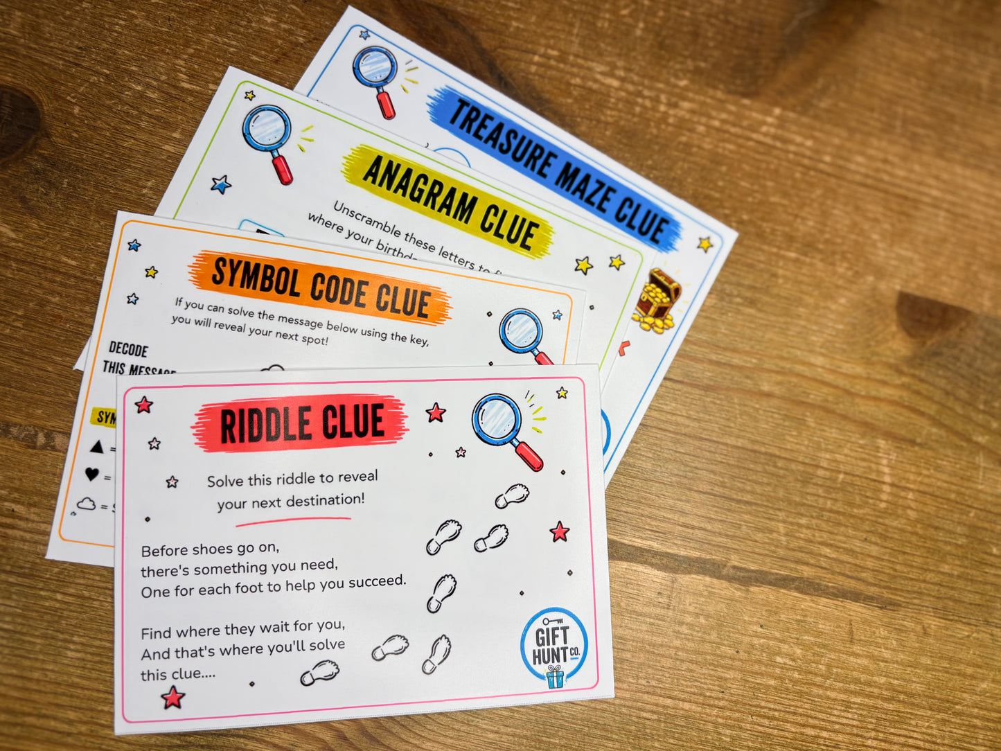 Three printed clue cards for a gift hunt game are arranged on a wooden tabletop. The cards read 'Treasure Maze Clue,' 'Anagram Clue,' and 'Symbol Code Clue,' with a front card titled 'Riddle Clue.