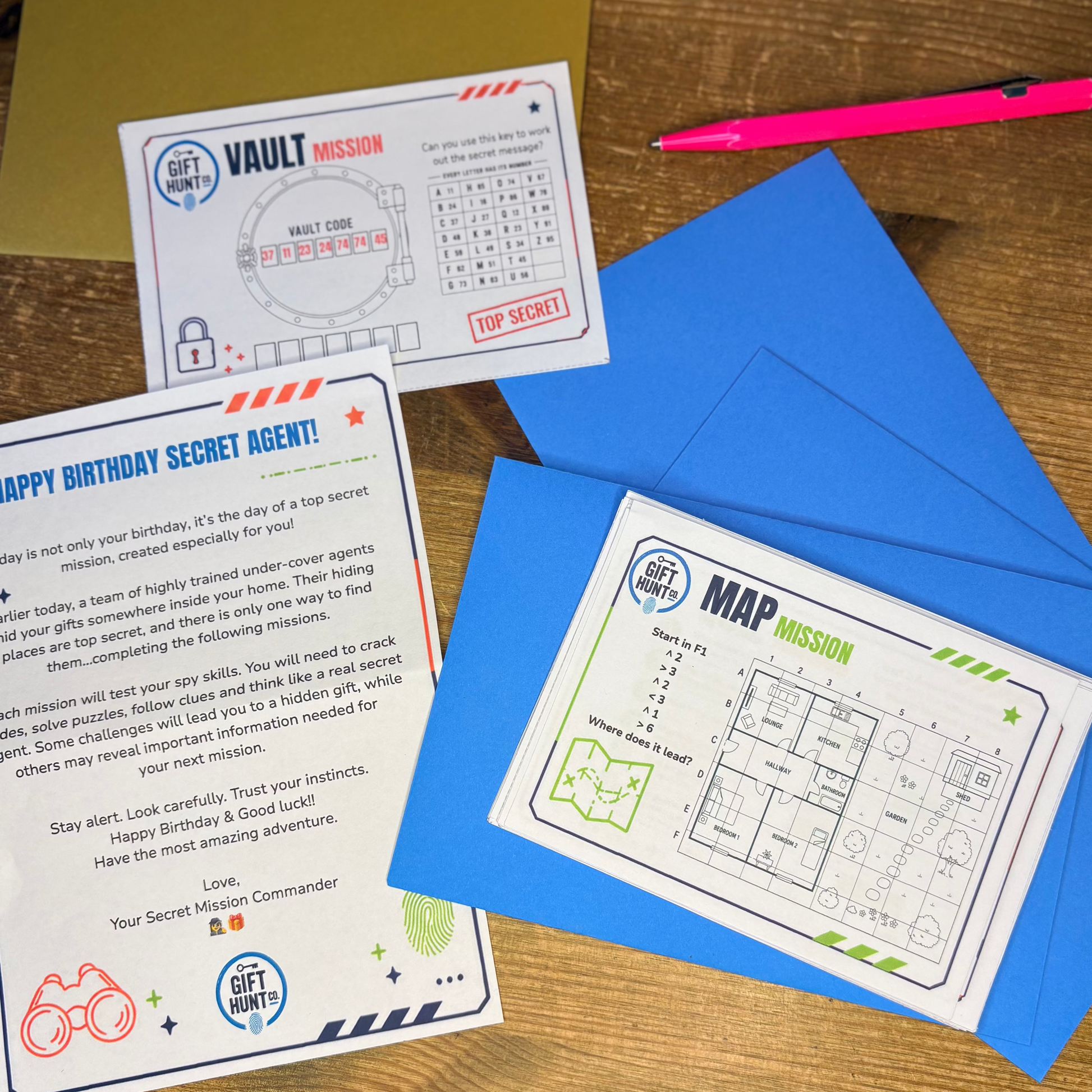 A set of printed secret agent themed cards and envelopes laid out on a wooden table, with a pink pen nearby. The cards feature mission prompts and grids under headings like Vault Mission and Happy Birthday Secret Agent.