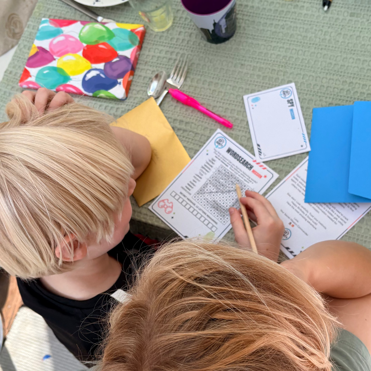 Two children sitting at a dining table, writing on a worksheet. The table is cluttered with school and household items including a purple mug, a colorful napkin, a blue notebook, and plates.